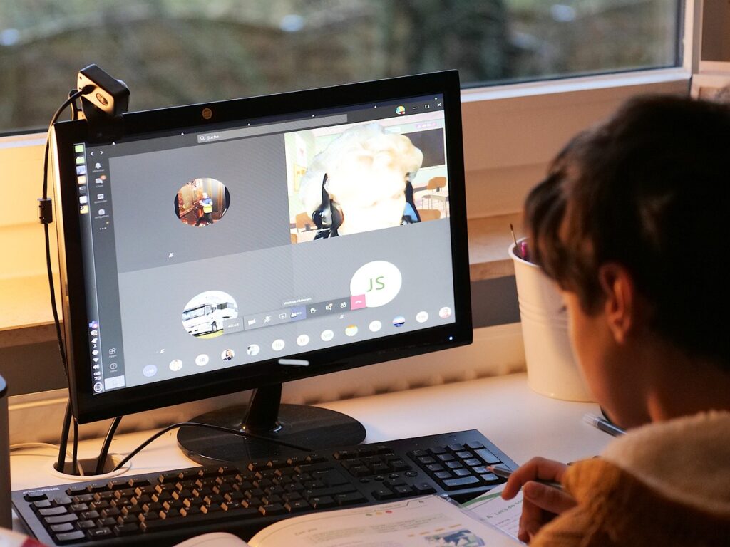 A photo of a student on a zoom call. The have a headset on and looking directly at the screen. On the screen, no ones camera is on, and you can see the student has text book sitting in front of them