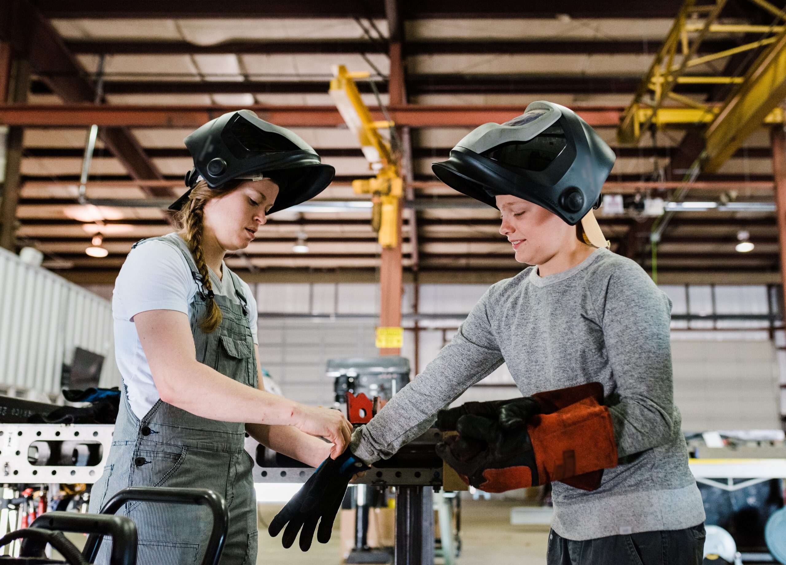 A woman welder helping another woman put on welding gloves.
