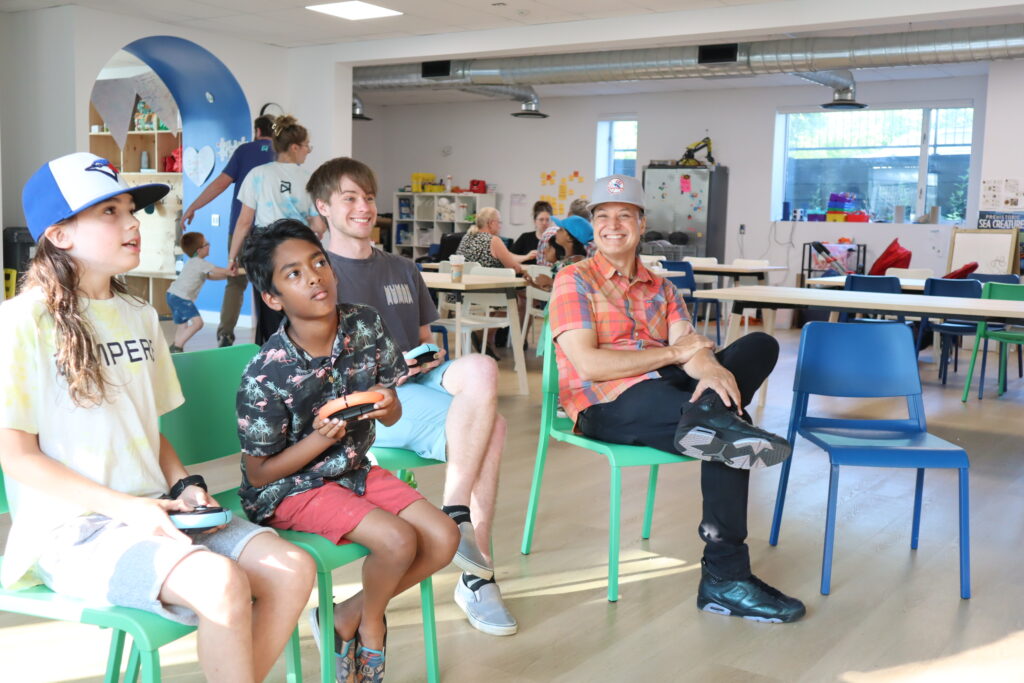 steam-fest-feature Three students on the left hand side are seen seated, with gaming controllers in their hands. An adult is seen seated to the right hand side, looking over at the students and smiling.