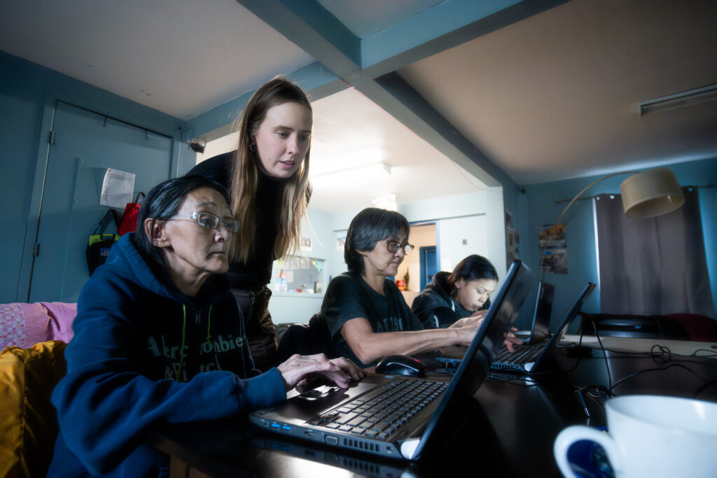 Three people are sitting down at a table and working on laptops. A STEAM educator is standing behind them looking at their screens.