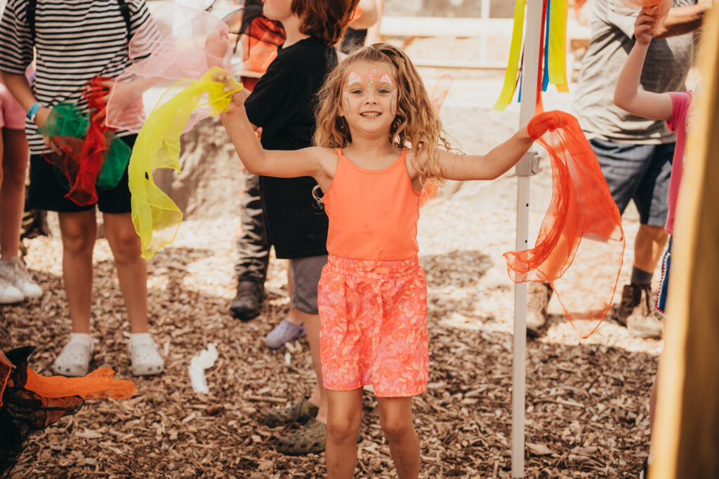 Young girl wearing orange clothes with her arms outstretched holding an orange streamer in one hand and a yellow streamer in her other. She's smiling with face paint on her face.