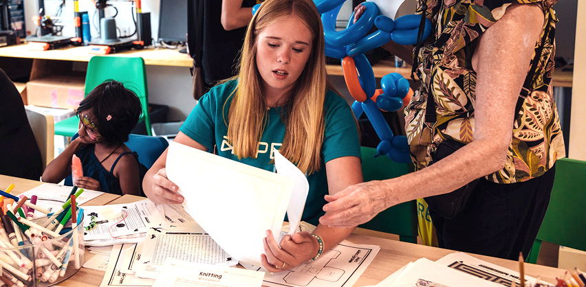 A STEAM educator is sitting down, showing activity sheets to a community member who is standing to her right side. 
