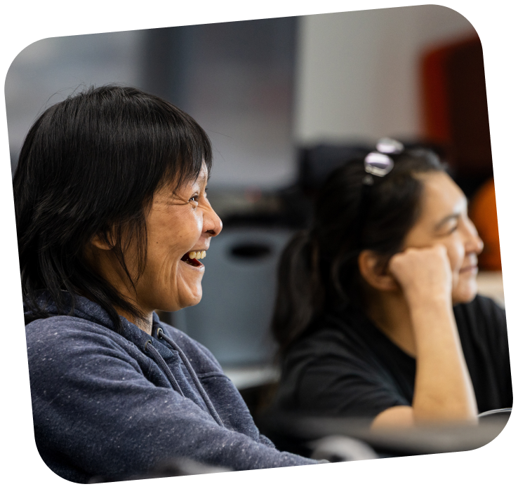 Two woman are in the frame, the one near the back is slightly out of focus and leaning on her hand, while the other woman is smiling.