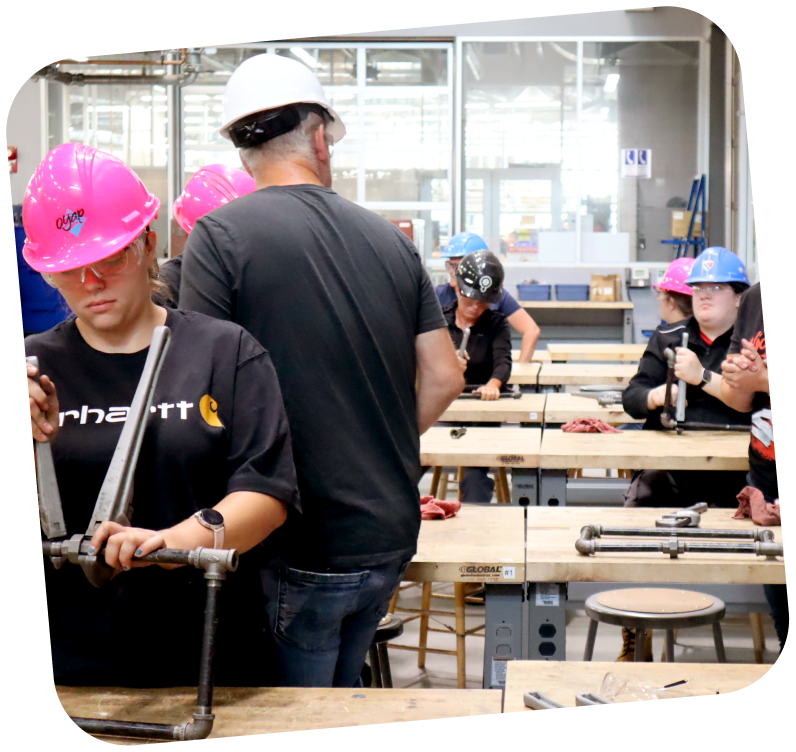 A classroom geared towards the trades is filled with students wearing hard hats and working on work tables.