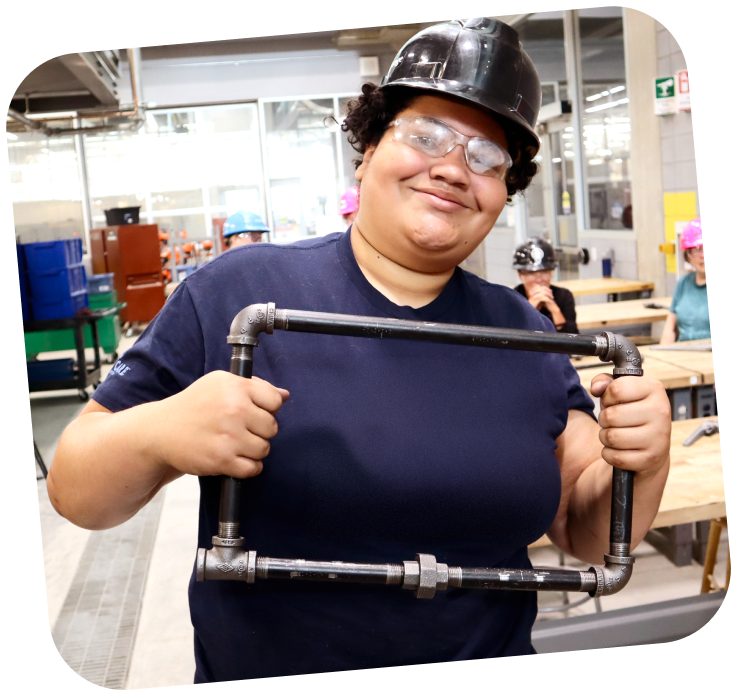 A student is holding a square prototype that looks to be welded together, wearing a hard hat. She is smiling at the camera.