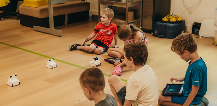 Children are sitting on the ground around a taped square. They are playing with little white robots that are driving around.