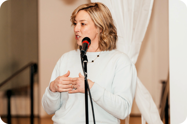 A woman with short hair is standing behind a mic, facing slightly to the left and is presenting.