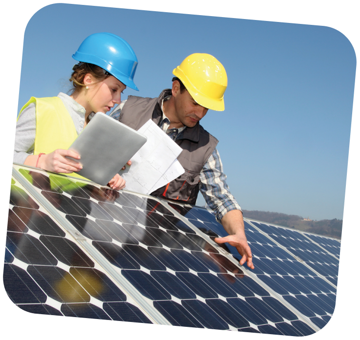 Two workers wearing hard hats and reflective vests are leaning over a solar panel. The sky is bright blue behind them.