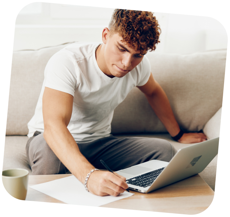 A young adult is sitting on a couch, with a laptop open in front of hum. He is writing something on a piece of paper that is located beside the laptop.