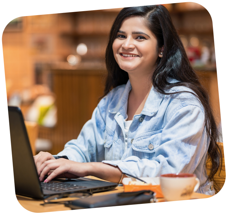 A young adult is sitting at a computer, looking over towards the camera and smiling.