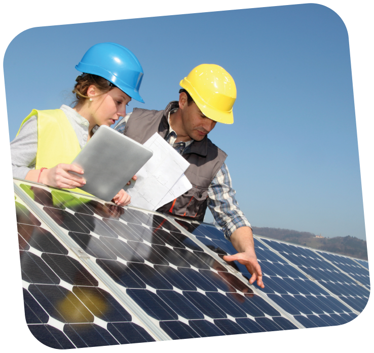Two workers wearing hard hats and reflective vests are leaning over a solar panel. The sky is bright blue behind them.