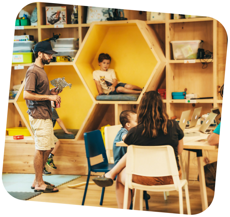 Families participating in activities taking place in the Makerspace. A child is sitting in a hexagon shaped seating area and the parents are around them.