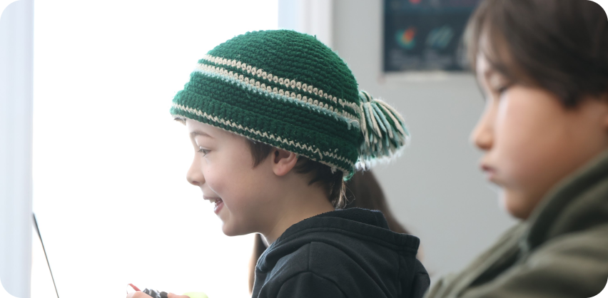 A young learner is sitting inside, wearing a pang hat. The learner is looking down slightly with a big smile on their face.