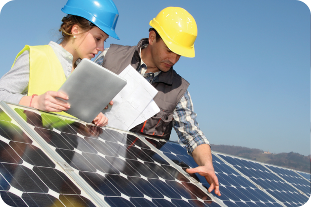 small-business-support-benefits-1 Two workers wearing hard hats and reflective vests are leaning over a solar panel. The sky is bright blue behind them.