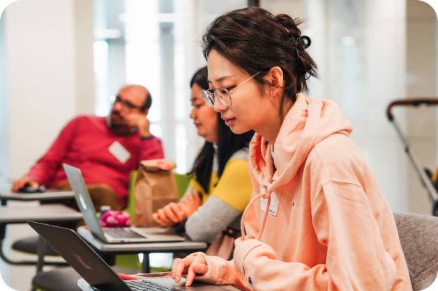 small-business-support-benefits-3-woman learning digital skills and AI A woman is looking downwards towards a computer. Two people are blurred behind her.
