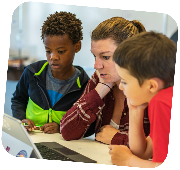 A STEAM educator is sitting between two learners, showing them something on a laptop. Both kids are interested and looking at the screen.