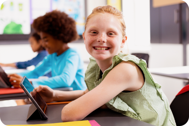 A younger learner sitting at a desk with an iPad in front of her, turned to her right and smiling at the camera.