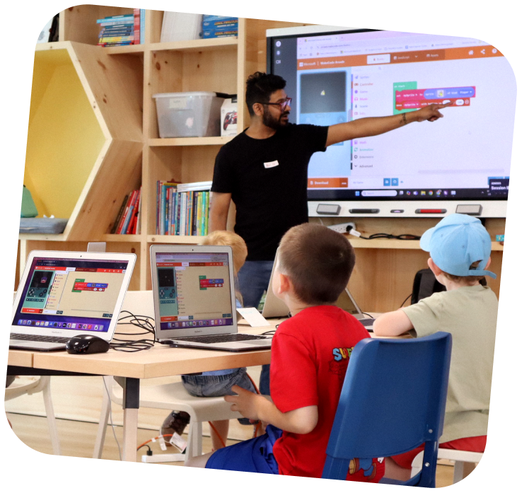 A STEAM educator is at the front of a Makerspace class, pointing at code blocks on a SmartBoard. The students are looking forward intently.