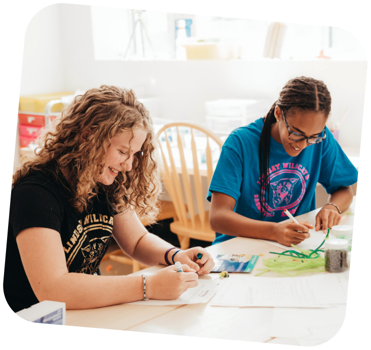Two teen girls working on an art project at the Lindsay Makerspace. They are sitting side by side at a table.