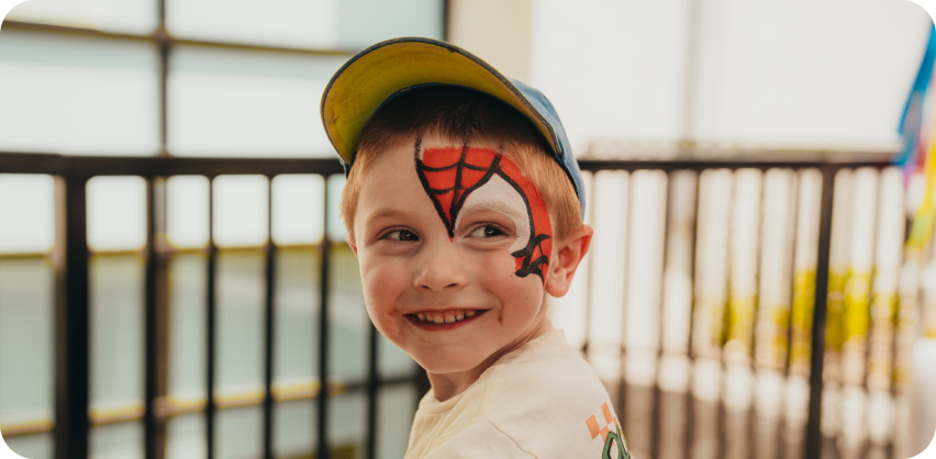 A young boy sitting outside. He is turned and smiling at the camera with a Spiderman face painting.