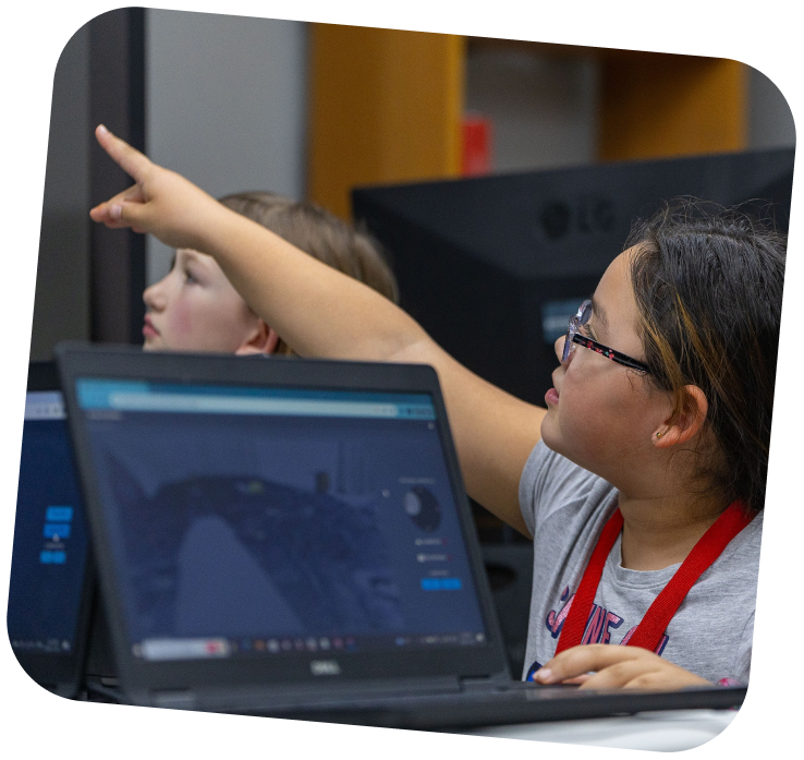 A younger learner is sitting beside an open laptop. She is looking forward towards another screen, and pointing at it.