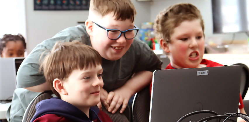 Two learners are sitting down and one is behind and leaned over them. They are all looking at a laptop.