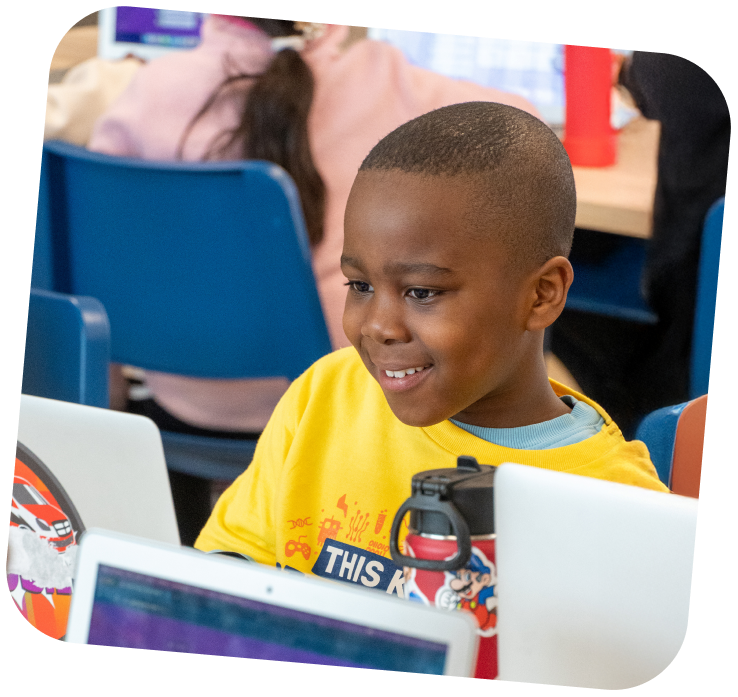 A young boy is sitting down at a table and smiling at the laptop sitting in front of him.