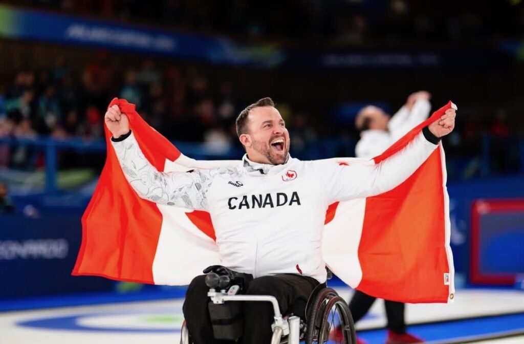 Jon Thurson, Paralympic gold medalist wearing a Team Canada shirt, proudly hoists a Canadian flag on a curling rink having just won at the Paralympics. 