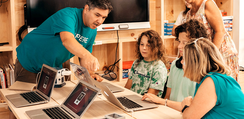makerspace-mentor-teaching-teens-digital-skills Two STEAM educators in blue ampere shirts sitting at a table with three students. They are looking and pointing towards one laptop screen.