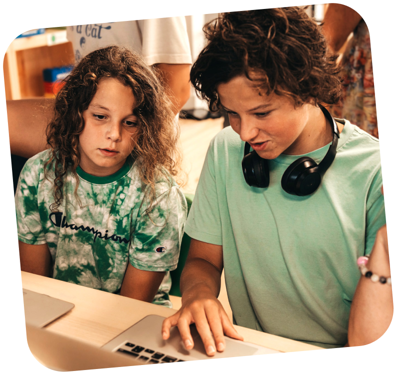 two-boys-learning-digital-skills Two boys are sitting at a table, the one closest to the camera has a hand on a track pad of a laptop while the other boy to his left looks at the computer screen.