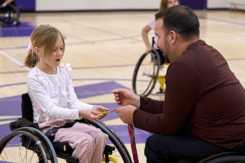Paralympian Jon Thurston, wearing a brown shirt and sitting in a wheelchair, show his Paralympic gold medal to a child in a wheelchair, who is looking inspired.