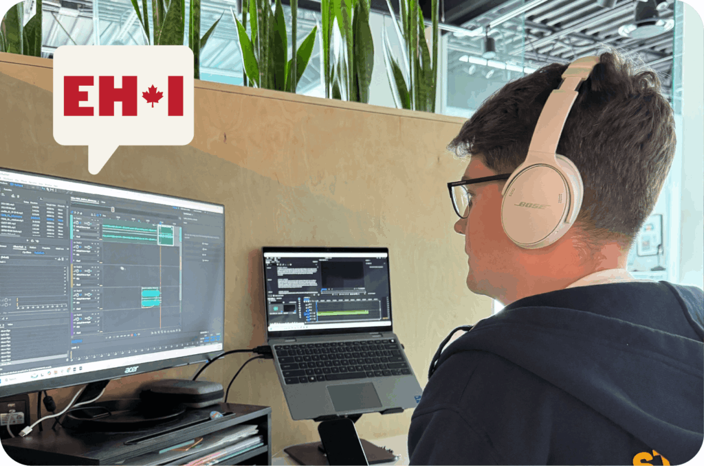 Eh I Podcast producer Zac Miller sits at a desk editing a new episode of the Eh I Podcast. There is a laptop on a stand and a monitor in front of Zac, who is wearing white headphones and a blue Ampere sweater.