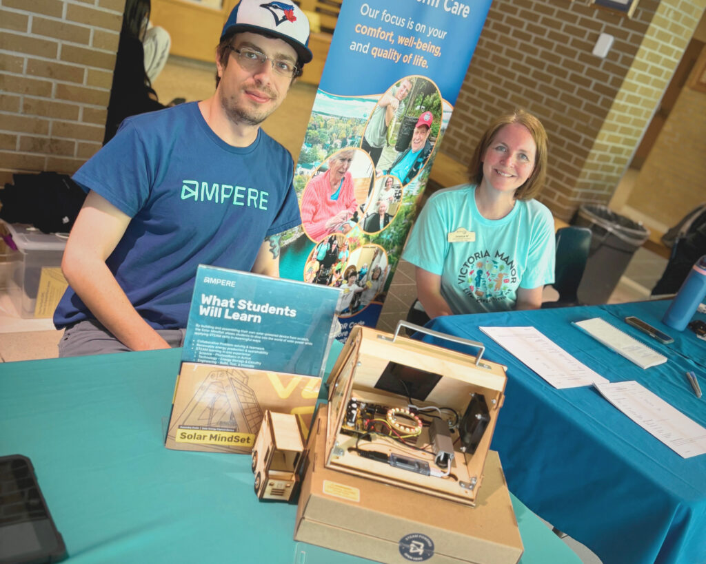 Two Ampere employees sit behind a table with a blue table cloth, looking at the camera. One is wearing a blue t-shirt with the Ampere logo on it, and the other a blue t-shirt with the LCVI logo on it. Various STEAM related items sit on top of the table.