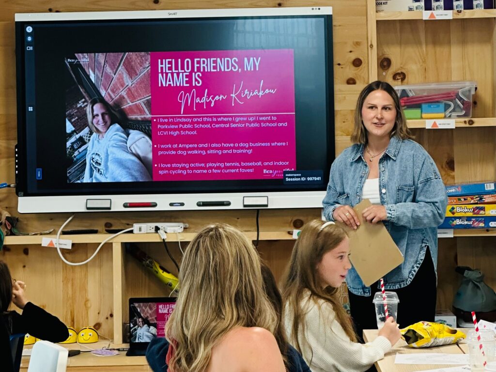 Madison, wearing a blue shirt, stands in front of group next to a Smart board, which features a presentation slide titled "hello friends, my name is Madison". Attendees are seating at tables in front of Madison.