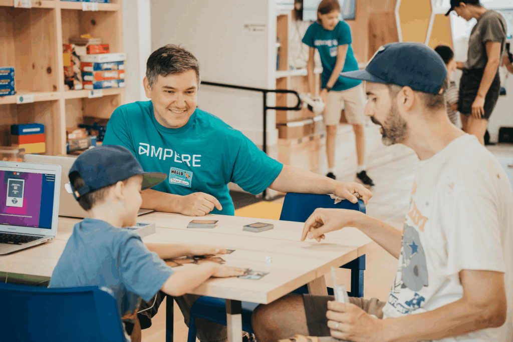 An instructor sits at a table in a classroom with a parent and a child. The instructor is wearing a teal t-shirt with an Ampere logo. The parent and child are wearing baseball caps and playing with cards on the table in front of them.