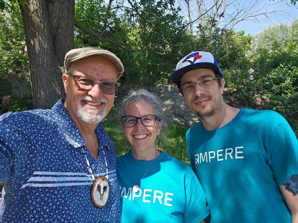fourth annual Pow Wow and Drum Circle in Gravenhurst three individuals pose for a photo outside, standing in front of green trees. They are all wearing blue shirts, two of which say AMPERE.