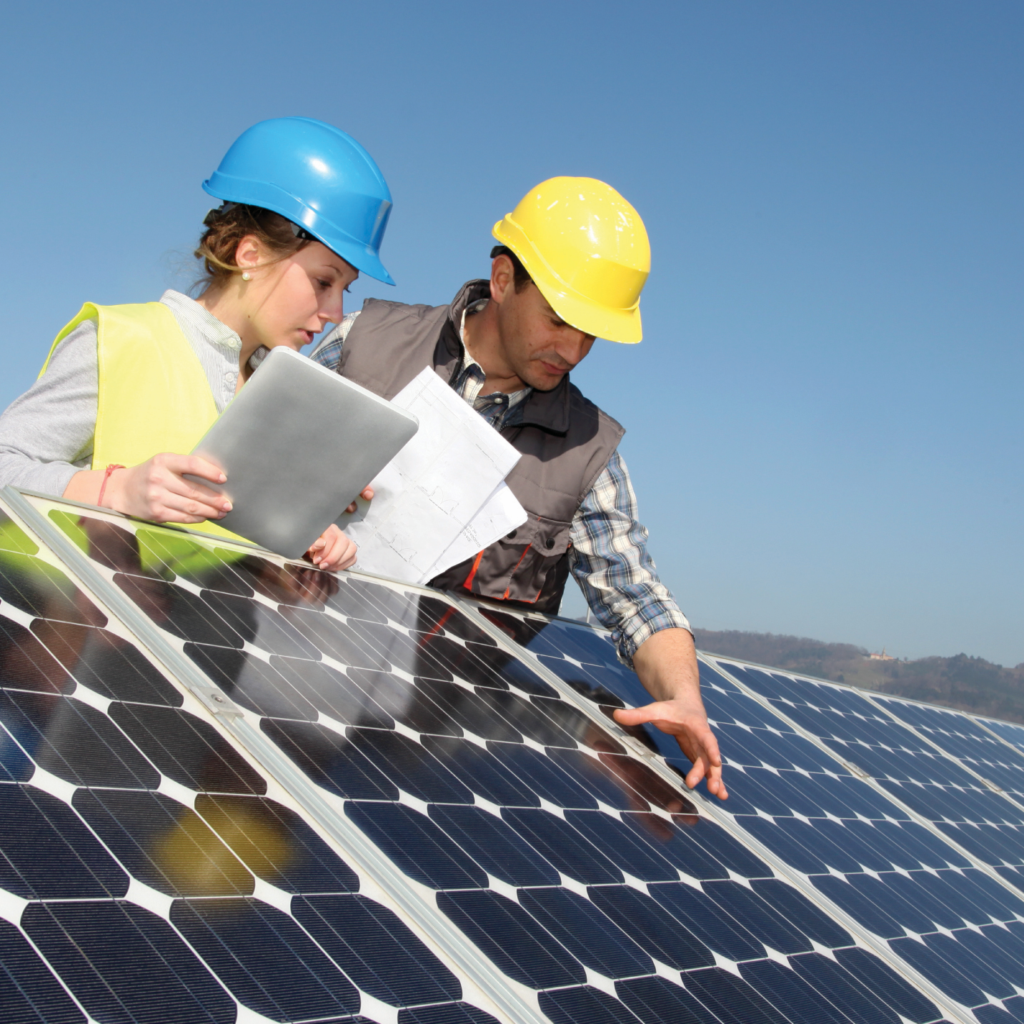 Two workers wearing hard hats and reflective vests are leaning over a solar panel. The sky is bright blue behind them.