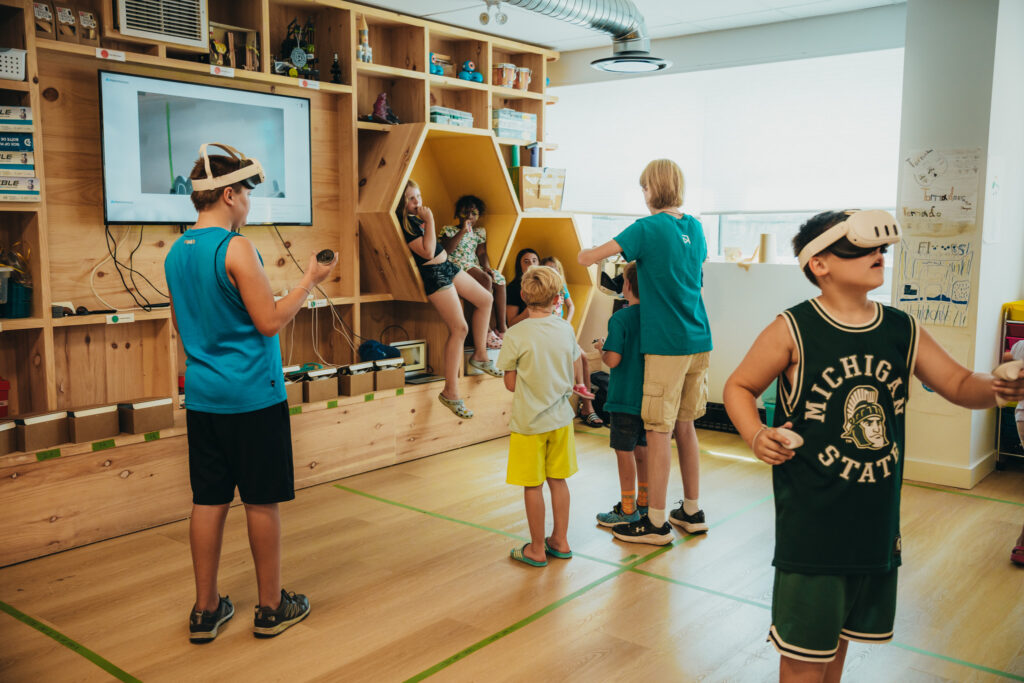 Students are standing in the makespace. Two of them have VR headsets on and are using the VR. The others are turned towards the SMARTBoard to view what they two play the VR are doing,
