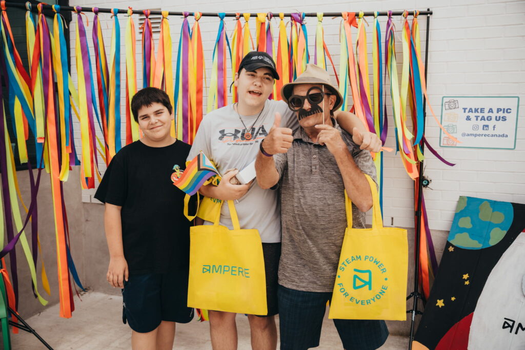 STEAM Fest 2025 A family stands in front of a photo booth with strings handing behind them. A gentleman with a beard holds a fake moustache in front of his face and has a thumbs up pointed at the camera. Two younger individuals stand next to him, smiling.