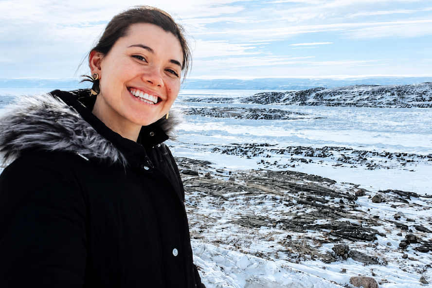 A woman stands in front of Arctic scenery. She is wearing a black coat with a fur collar, looking at the camera and smiling. Her hair is black. The sky is blue, but there are clouds throughout.