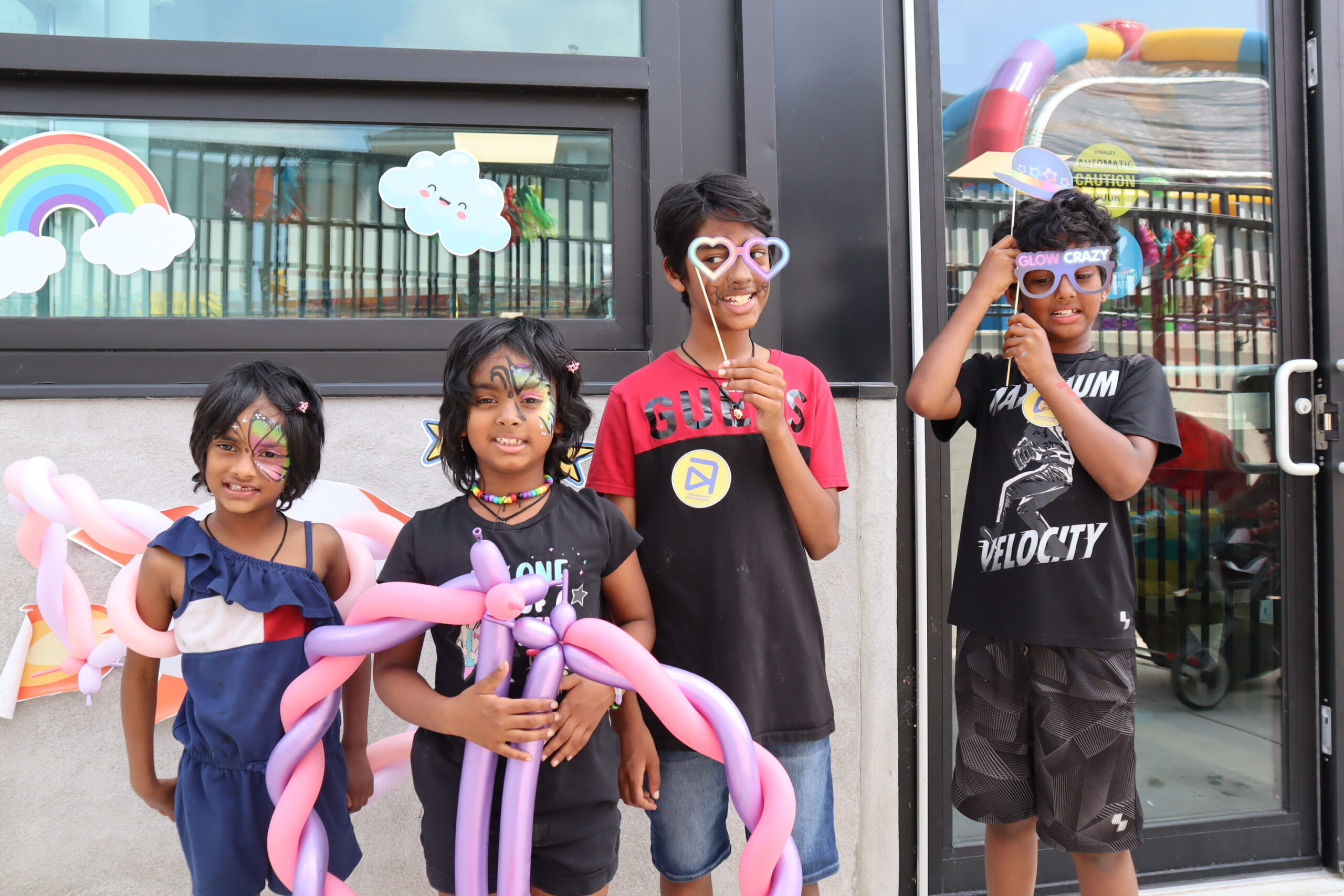 Four children stand at a street festival wearing props like glasses and balloon animals.