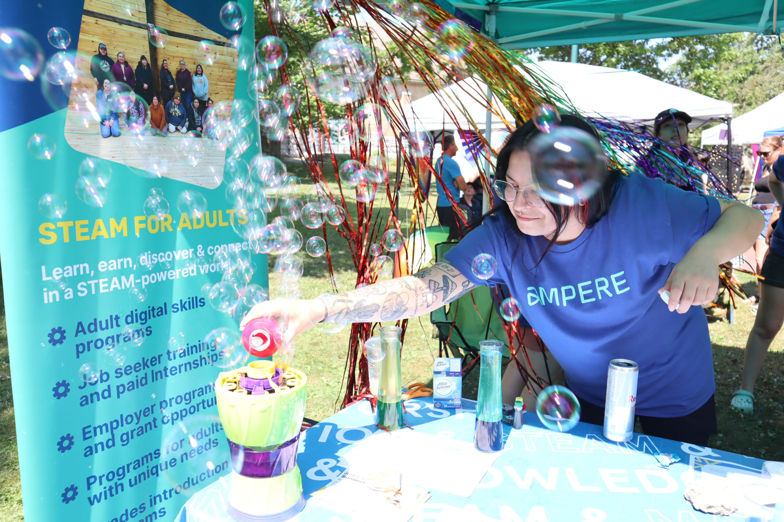 A woman pours liquid into a bubble machine at a blue table with a blue banner behind her that says Ampere. She is wearing glasses and a purple t-shirt that says Ampere.
