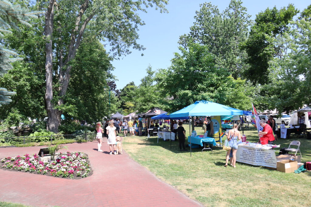 a large tent is set up in the middle of a green park. Trees have all their leaves in the middle of summer. There are people walking along the path wearing red t-shirts.