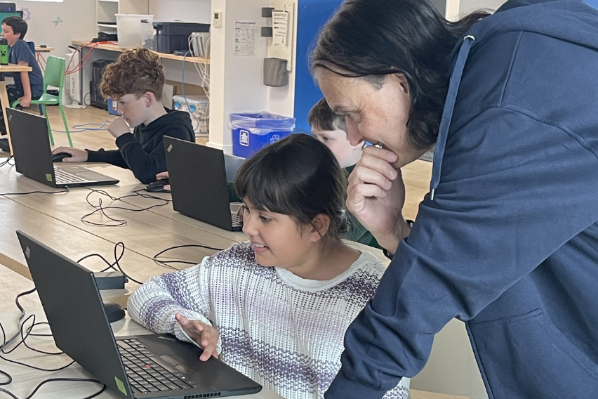 STEAM School Recap 1 Kevin, wearing a blue hoodie, instructs a child wearing a grey sweater how to use a computer. Other children are using computers in the background.