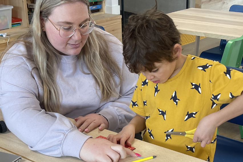 Mel, wearing glasses and a grey sweater, teaches a child in a yellow shirt how to use a ruler.