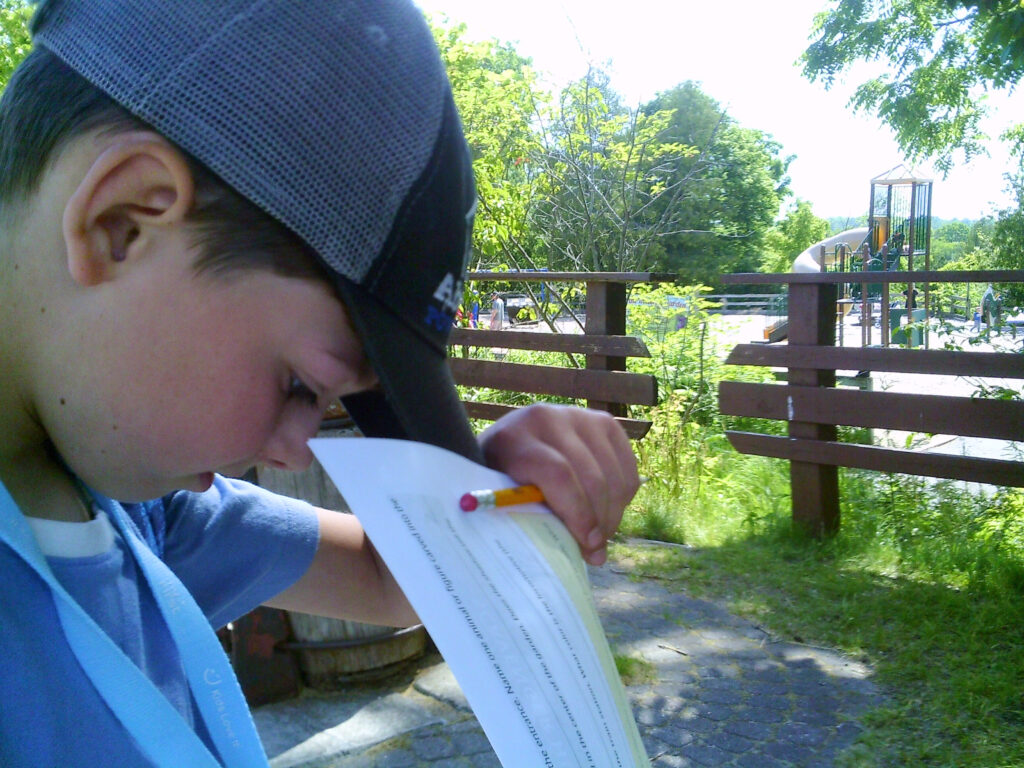 A student is holding a paper and pencil, writing notes at the Zoo about the animals. He has a grey baseball cap on his head and is wearing a blue t-shirt. 