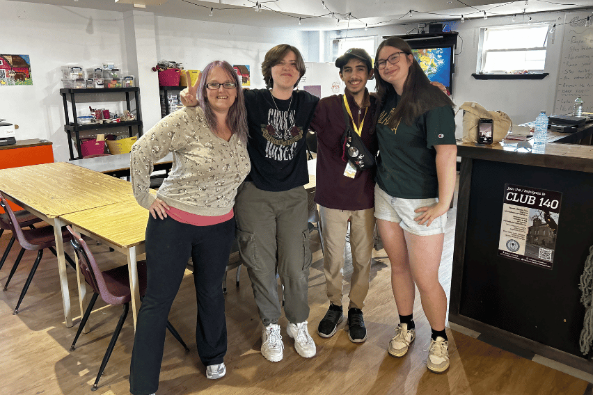 Bathurst Makerspace Team 4 individuals pose for a photo in a classroom makerspace environment. Desks are visible behind them. They ave their arms over each others shoulders and are all smiling.