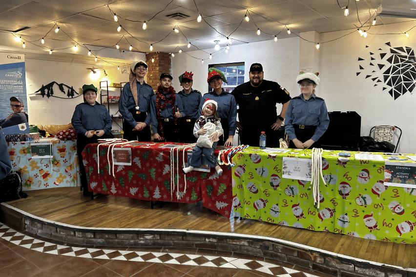 Bathurst Makerspace Christmas A group of adults and children stand wearing festive hats behind festive tables with green and red tablecloths.
