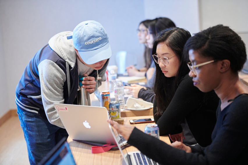 An instructor shows two students ow to use a program on a laptop that is sitting between them. The instructor is wearing a blue baseball cap. Two more students sit behind the group doing their own work.