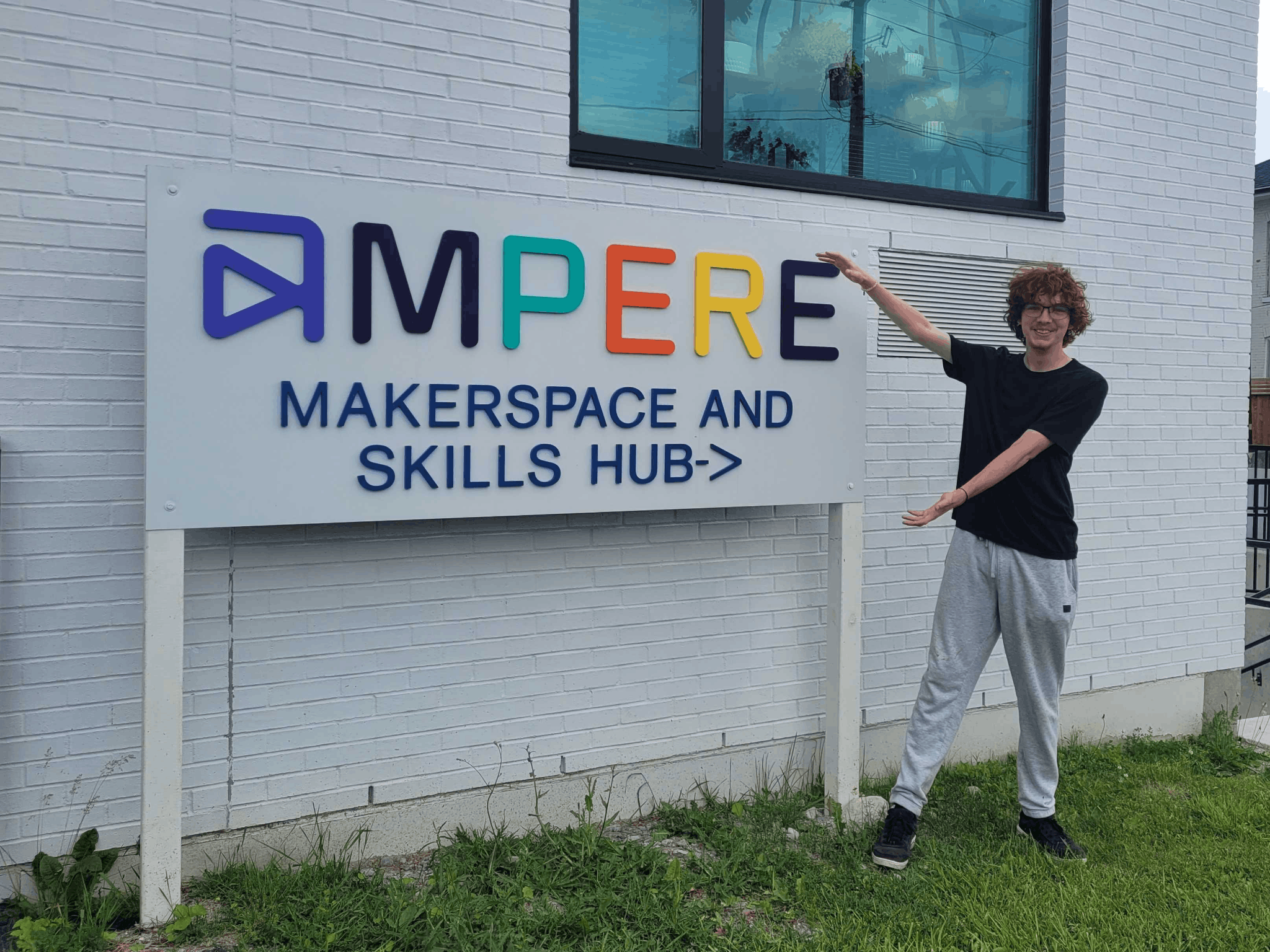 Zach Callan stands in front of the Ampere Kawartha Lakes Makerspace & Skills Hub sign in Lindsay, ON. He's wearing a black shirt and grey sweatpants.